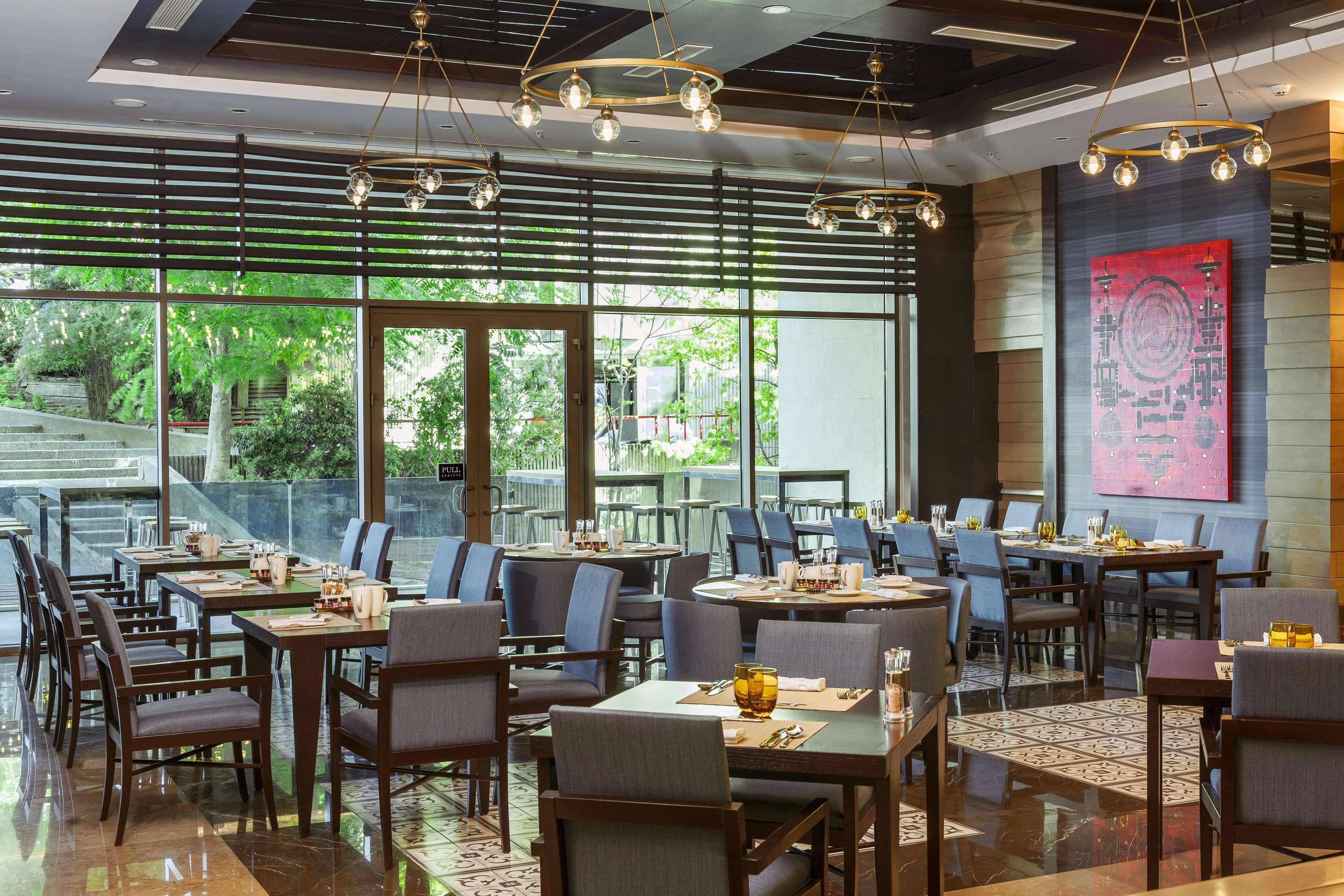 Radisson Blu Hotel, Istanbul Sisli Exterior photo The photo shows a modern restaurant interior. The space features several tables and chairs arranged neatly, with a mix of wooden and upholstered furniture. Large windows let in natural light and offer a view of greenery outside. The decor has a conte