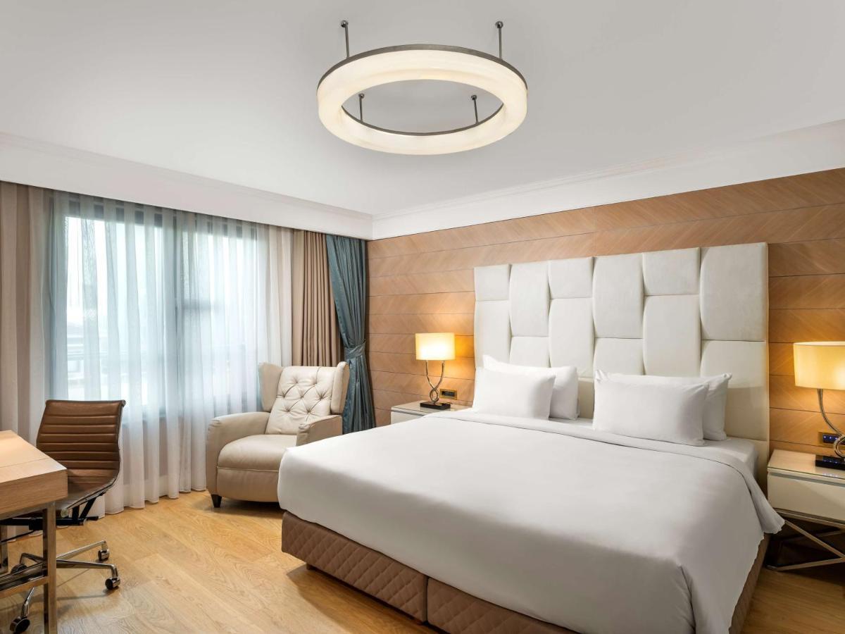 Radisson Blu Hotel, Istanbul Sisli Exterior photo The photo shows a modern hotel room featuring a large bed with a light-colored headboard. There are two bedside lamps on either side of the bed. To one side, there is a comfortable armchair and a small wooden desk with a rolling chair. The room has l
