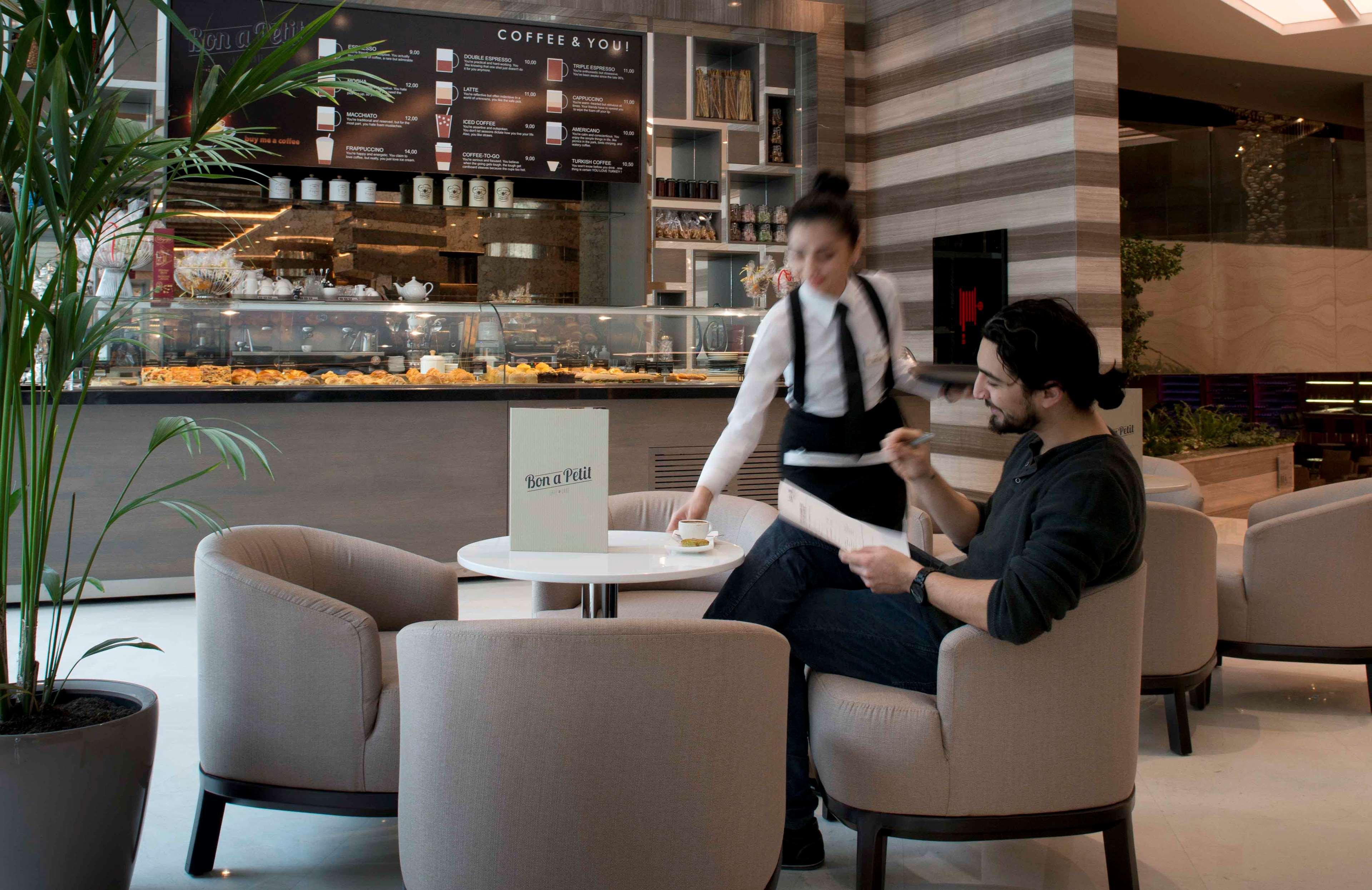 Radisson Blu Hotel, Istanbul Sisli Exterior photo The photo shows a modern cafe or coffee shop setting. In the foreground, a man is seated at a small round table, looking at a menu or paper in his hand. He is dressed casually in a dark sweater. A waitress, wearing a white shirt and black apron, is s
