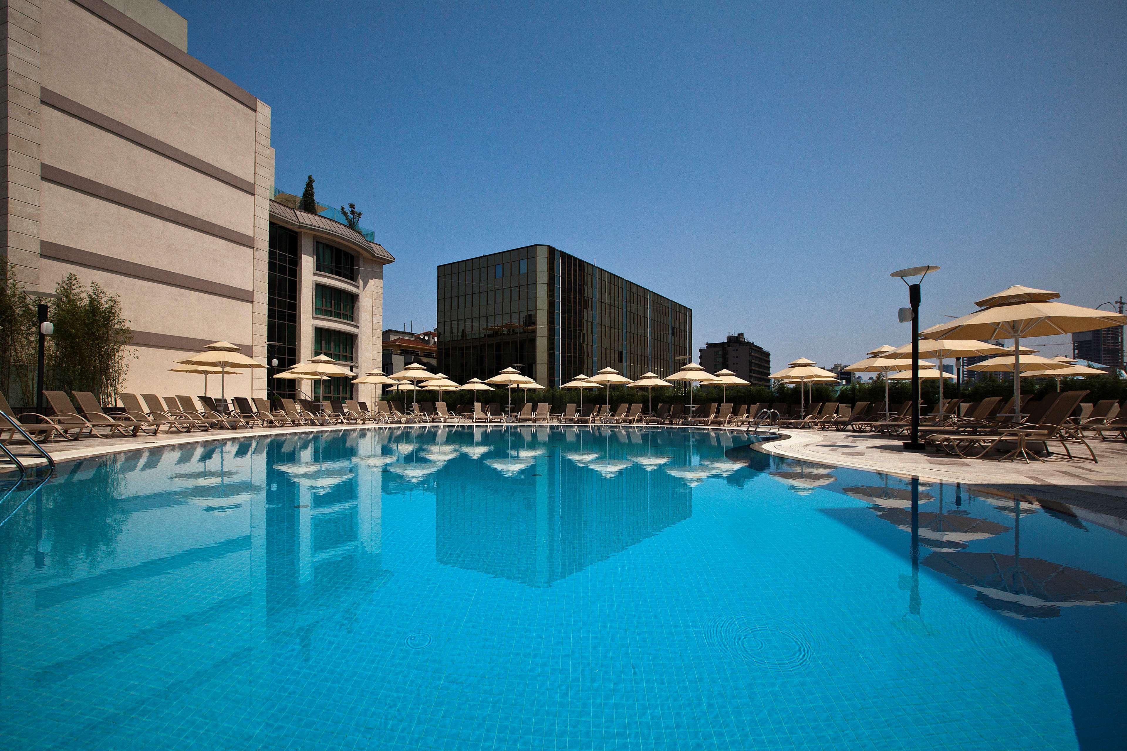 Radisson Blu Hotel, Istanbul Sisli Exterior photo The photo shows a serene swimming pool surrounded by lounge chairs and umbrellas. The pool has a clear, inviting blue water, reflecting the bright blue sky above. In the background, there are two buildings: one appears to be modern with large windows