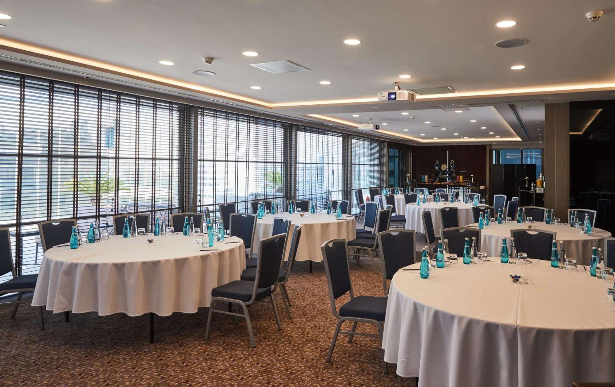 Radisson Blu Hotel, Istanbul Sisli Exterior photo The photo shows a conference or meeting room set up for an event. There are several round tables covered with white tablecloths, each accompanied by chairs. Atop the tables, there are glass bottles of water. The room features large windows with blind